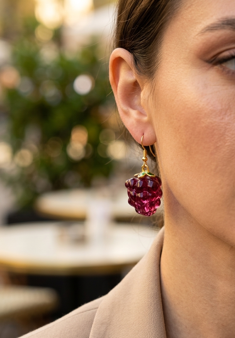 Arete con dije de frambuesa de resina roja translúcida y detalles dorados, usado por una mujer con blusa de lino con luz natural.