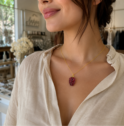 Collar con dije de frambuesa de resina roja translúcida y detalles dorados, usado por una mujer con blusa de lino en una boutique iluminada con luz natural.
