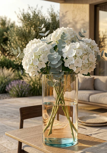 Florero cilindro de vidrio transparente con un arreglo de hortensias blancas y eucalipto como centro de mesa en una terraza al aire libre