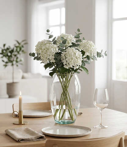 Florero de vidrio Medellín transparente 16x25 cm con hortensias blancas y eucalipto sobre una mesa de comedor en un espacio moderno con decoración minimalista.