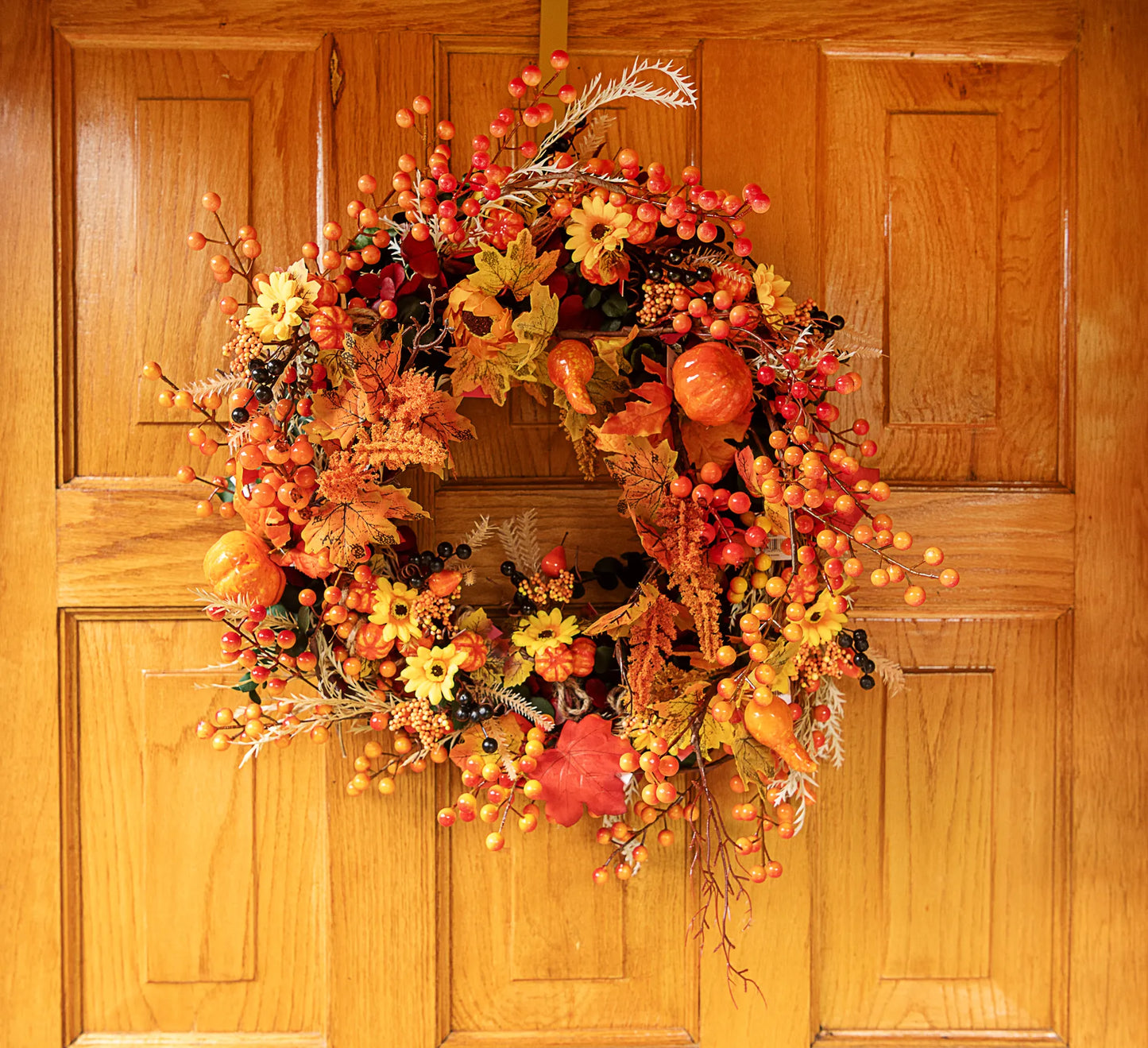 Corona decorativa de otoño elaborada con picks artificiales de bayas, hojas de maple, calabazas y flores sobre una puerta de madera.