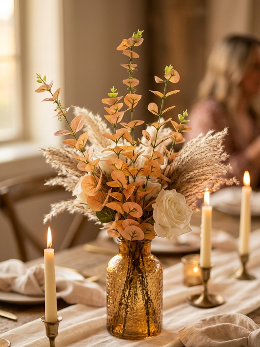 Rama de eucalipto artificial en tono dorado colocada en un florero de vidrio ámbar como centro de mesa para boda, combinada con rosas blancas y pampas, ideal para decoración de eventos elegantes.