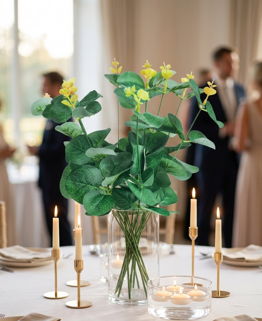 Rama de eucalipto artificial verde con brotes amarillos colocada en un florero de vidrio como centro de mesa para boda o evento, ideal para decoración elegante y arreglos florales.